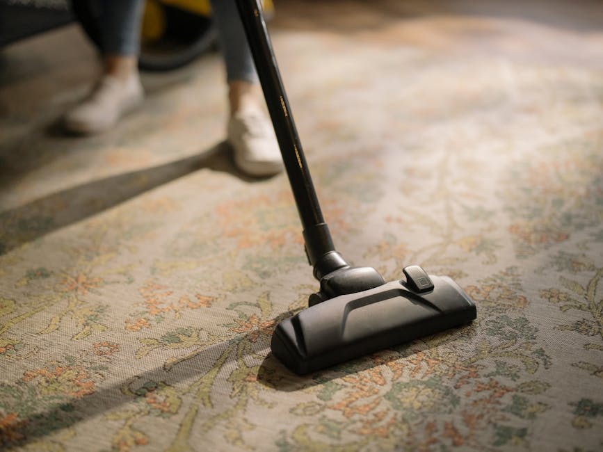 Close-up of a black vacuum cleaner attachment being used to deep clean a patterned area rug with floral motifs in a residential space. The vacuum head is flat against the beige and pastel-colored carpet, and part of the vacuum wand extends upward, held by a person's hand, with the person partially visible in the background wearing casual shoes. The room is well-lit, highlighting the cleanliness of the surface, and the scene reflects professional surface cleaning and maintenance carried out by [COMPANY_NAME], as part of domestic cleaning services in Kingston upon Thames.