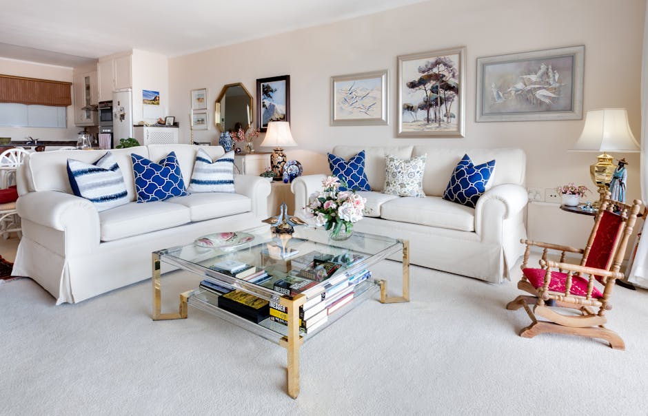 A bright, well-maintained living room with cream-colored carpet flooring featuring a glass-top coffee table with a gold frame, topped with a floral arrangement and magazines stored underneath. Two white upholstered sofas with blue and white patterned cushions are positioned against the wall, which is adorned with three framed landscape artworks. To the right, a small wooden rocking chair with red upholstery is placed next to a side table holding a decorative lamp and a small potted plant. The room is illuminated by natural daylight and a table lamp, creating a warm, inviting atmosphere suitable for domestic cleaning and surface sanitisation activities. In the background, part of an open kitchen with wooden cabinets and a white countertop is visible.