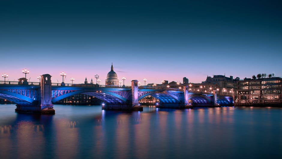 A wide view of Westminster Bridge illuminated with blue and pink lighting, spanning the River Thames during twilight, with the historic St. Paul's Cathedral domed roof visible in the background under a darkening sky. The bridge's detailed ironwork and street lamps are clearly visible, reflecting softly in the calm water below. The scene conveys a sense of cityscape cleanliness and well-maintained infrastructure near central London, aligning with the themes of surface cleaning and urban sanitation, as featured on carpetcleaningkingstonuponthames.com.
