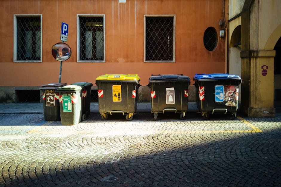 A blue recycling bin positioned on a flat surface next to a white commercial delivery truck with closed, plain side panels, situated outdoors. The bin appears to be made of plastic, with a lid and whe