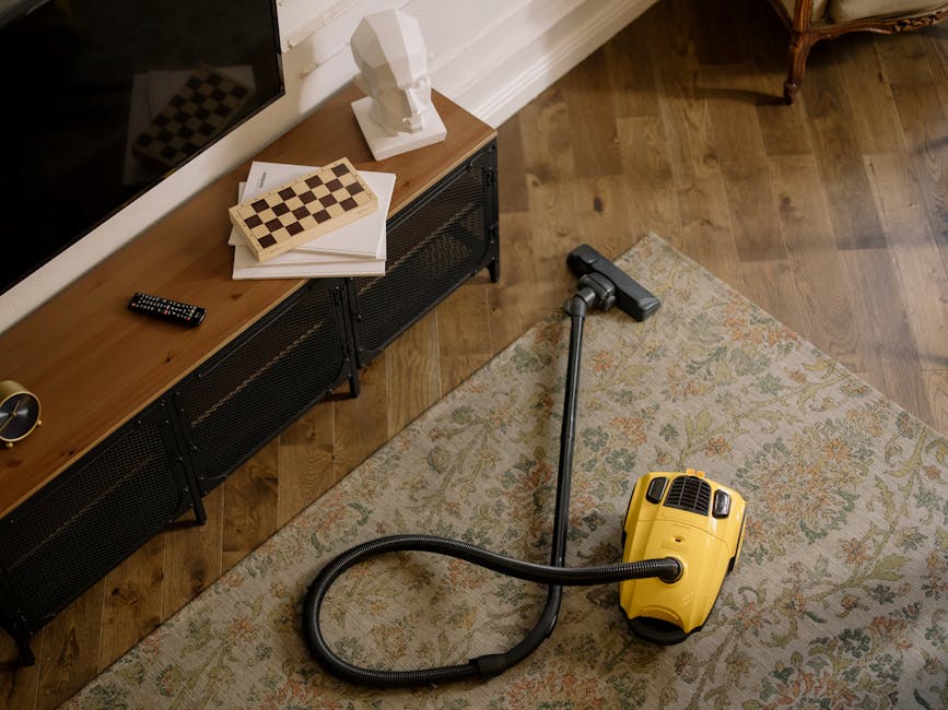 A living room during surface cleaning, featuring a white vacuum cleaner with a black hose on a wooden floor next to a beige sofa adorned with blue and white cushions. A man with gray hair and a young 